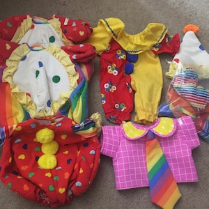 May include: A set of five colorful clown costumes for dolls. The costumes include a red and yellow jumpsuit with a white collar, a red and white striped shirt with a rainbow tie, a red and white striped hat, a red and white striped shirt with a rainbow tie, and a red and white striped shirt with a rainbow tie.