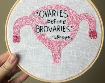 May include: Hand-stitched embroidery hoop art featuring a pink uterus design. The text reads "Ovaries before Brovaries" in black thread, with "-L. Knope" below. The artwork is set in a wooden embroidery hoop.