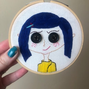 Op de afbeelding: Borduurwerk in een hoepel met een cartoonmeisje met blauw haar, zwarte knoopjesogen en een geel shirt.