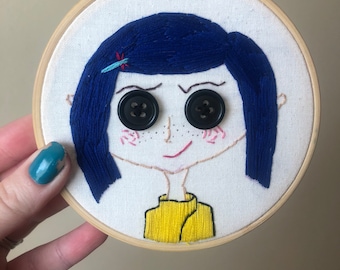 May include: Embroidered hoop art featuring a cartoon girl with blue hair, black button eyes, and a yellow shirt.
