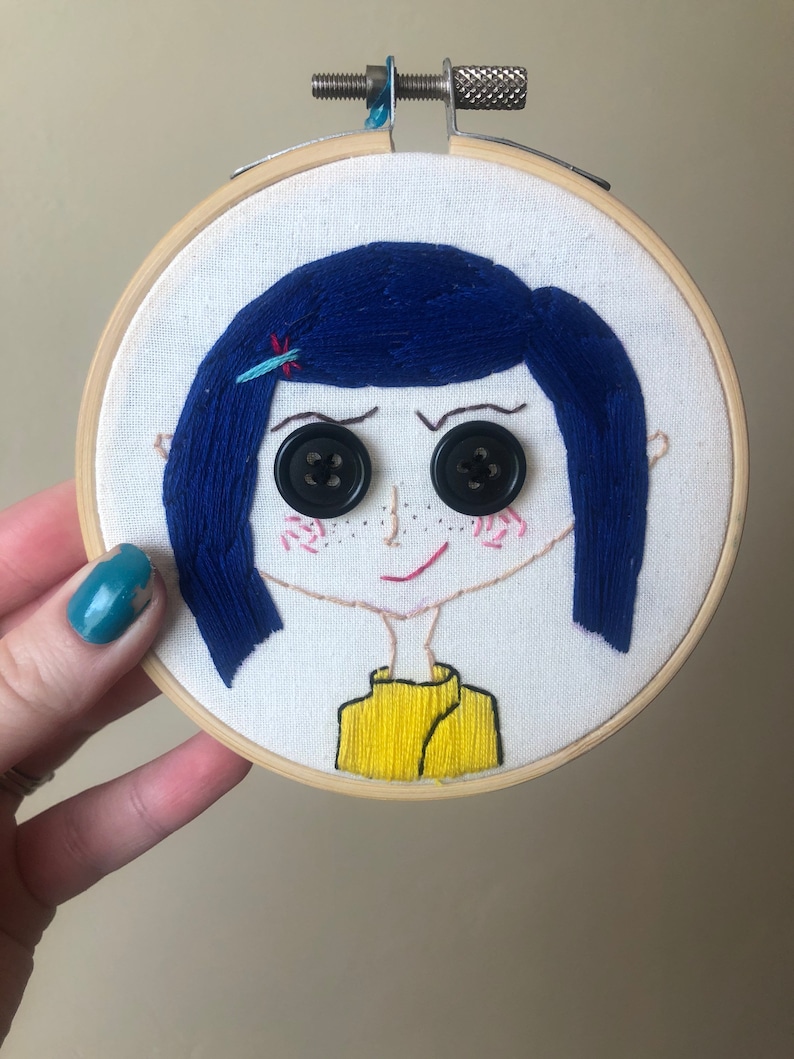 Op de afbeelding: Borduurwerk in een hoepel met een cartoonmeisje met blauw haar, zwarte knoopjesogen en een geel shirt.