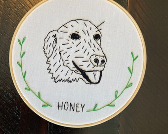 May include: Embroidered dog portrait in a wooden hoop. The dog's face is rendered in black thread with a minimalist style. The name "HONEY" is stitched below the dog. Green embroidered leaves frame the design.
