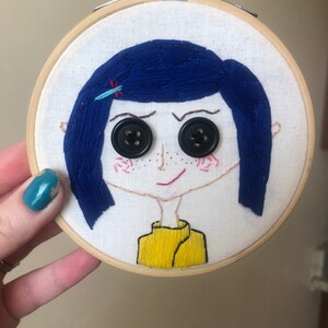 Op de afbeelding: Borduurkunst in een hoepel met een cartoonmeisje met blauw haar, knoopjesogen en een geel shirt.