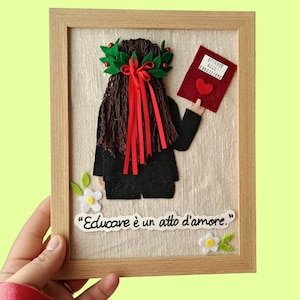 Puede incluir: Una obra de arte enmarcada de fieltro que representa a una persona graduada sosteniendo un libro. La figura tiene una corona de laurel y cintas rojas. Se incluye el texto "Educare è un atto d'amore". El marco es de madera.
