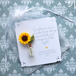 May include: A white card with a yellow sunflower and a small glass bottle filled with white beads. The card has a message about sunflowers and is inside a sheer white bag. The background is a patterned blue fabric.