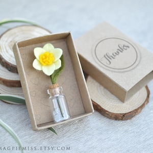 May include: A small, yellow felt daffodil flower in a glass vial with white stones, inside a brown cardboard box with the word "Thanks" printed on the lid.