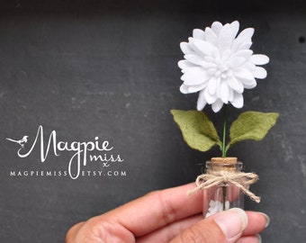 May include: A handmade white felt flower with green felt leaves and a cork base, displayed in a small glass bottle. The bottle is tied with twine and contains small white objects. The background is a dark gray chalkboard.