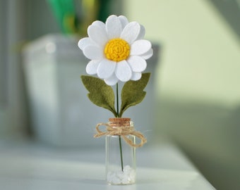 May include: A single white felt daisy with a yellow center is displayed in a small glass jar with a cork top. The jar is filled with white decorative stones and tied with twine.
