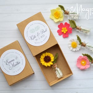 Pu&ograve; includere: Scatole regalo marroni con decorazioni floreali ed etichette personalizzate. Ogni scatola contiene un fiore in miniatura e una piccola bottiglia di vetro piena di perline bianche. Le etichette recitano "Grazie per aver festeggiato con noi oggi" e includono nomi e date.