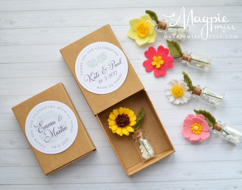Pu&ograve; includere: Scatole regalo marroni con decorazioni floreali ed etichette personalizzate. Ogni scatola contiene un fiore in miniatura e una piccola bottiglia di vetro piena di perline bianche. Le etichette recitano "Grazie per aver festeggiato con noi oggi" e includono nomi e date.