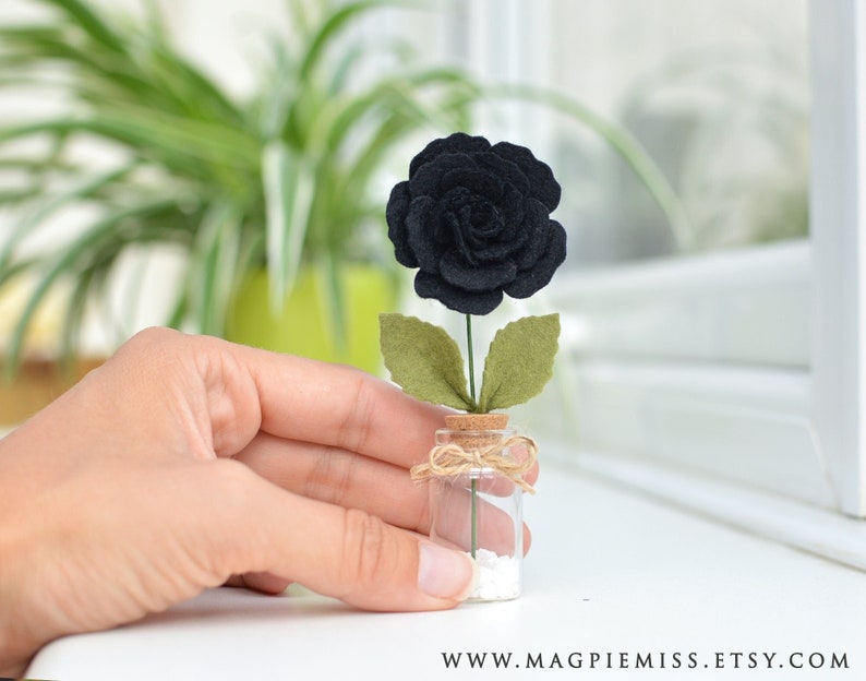 May include: A single black felt rose with green felt leaves in a small glass jar with a cork top and twine tied around the cork. White decorative stones are at the bottom of the jar.