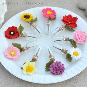 Pu&ograve; includere: Un piatto bianco mostra piccole bottiglie di vetro con tappi di sughero, ciascuna con uno stelo e sormontata da un fiore di feltro. I fiori sono in vari colori, tra cui rosso, rosa, giallo e viola, con foglie di feltro verde.