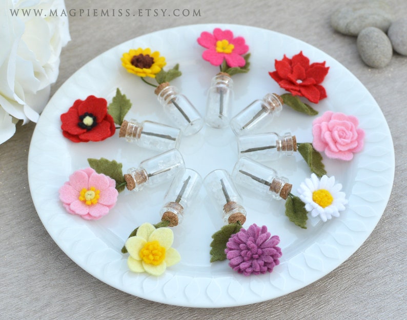 Pu&ograve; includere: Un piatto bianco mostra piccole bottiglie di vetro con tappi di sughero, ciascuna con uno stelo e sormontata da un fiore di feltro. I fiori sono in vari colori, tra cui rosso, rosa, giallo e viola, con foglie di feltro verde.