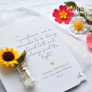May include: A white card with a sunflower design and the text "Sunflowers are a reminder to be strong, stand tall and always find the light." A small glass bottle with a sunflower and a small white flower is attached to the card. Other felt flowers are in the background.