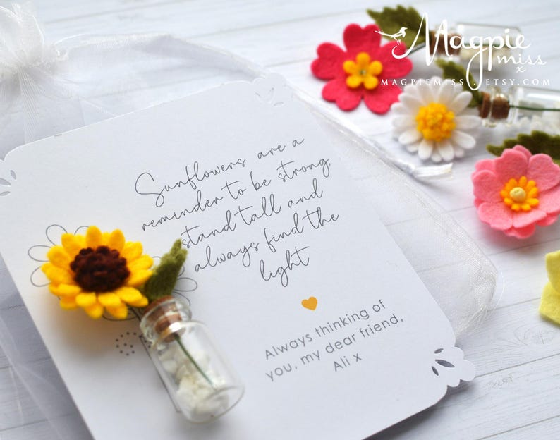 May include: A white card with a sunflower design and the text "Sunflowers are a reminder to be strong, stand tall and always find the light." A small glass bottle with a sunflower and a small white flower is attached to the card. Other felt flowers are in the background.