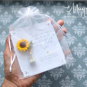 May include: A small, clear glass vial with a miniature yellow sunflower and a white card inside a sheer white gift bag. The card has text that reads "Sunflowers are a reminder to be strong..." and "Always thinking of you, my dear friend, Alix."