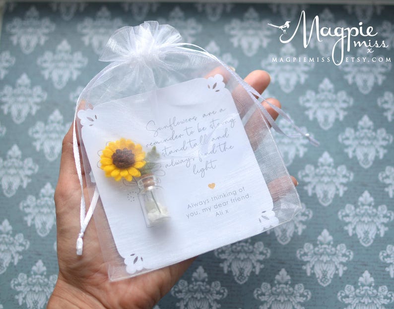 May include: A small, clear glass vial with a miniature yellow sunflower and a white card inside a sheer white gift bag. The card has text that reads "Sunflowers are a reminder to be strong..." and "Always thinking of you, my dear friend, Alix."