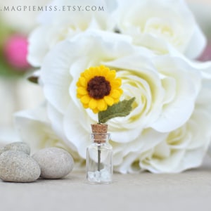 Pu&ograve; includere: Una piccola bottiglia di vetro con un tappo di sughero contenente un piccolo girasole di feltro. Il girasole ha petali gialli e un centro marrone, con una foglia verde di feltro. Sullo sfondo sono visibili rose bianche e piccole pietre grigie.