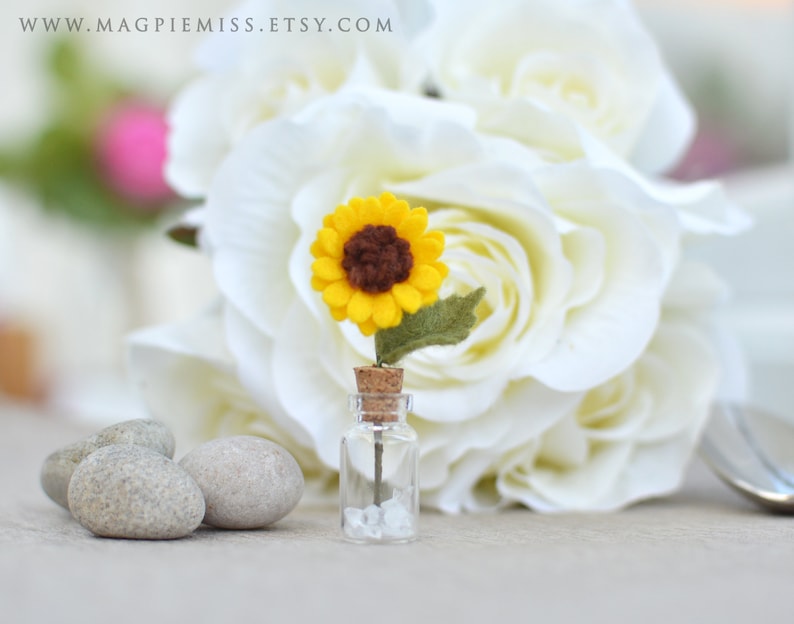 Pu&ograve; includere: Una piccola bottiglia di vetro con un tappo di sughero contenente un piccolo girasole di feltro. Il girasole ha petali gialli e un centro marrone, con una foglia verde di feltro. Sullo sfondo sono visibili rose bianche e piccole pietre grigie.