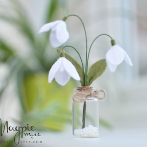 K&ouml;nnte beinhalten: Eine kleine Glasflasche mit drei wei&szlig;en Schneegl&ouml;ckchen aus Filz, gr&uuml;nen Stielen und Bl&auml;ttern. Die Flasche ist mit Bindfaden gebunden und mit kleinen wei&szlig;en Steinen gef&uuml;llt. Der Hintergrund ist verschwommen, mit Gr&uuml;n- und Wei&szlig;t&ouml;nen.