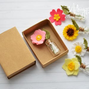 Pu&ograve; includere: Una scatola artigianale con un fiore di feltro rosa e una piccola bottiglia di vetro piena di perline bianche. Diversi fiori di feltro colorati e bottiglie di vetro con perline bianche sono disposti su una superficie di legno bianco.