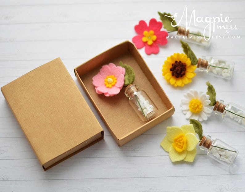 Pu&ograve; includere: Una scatola artigianale con un fiore di feltro rosa e una piccola bottiglia di vetro piena di perline bianche. Diversi fiori di feltro colorati e bottiglie di vetro con perline bianche sono disposti su una superficie di legno bianco.