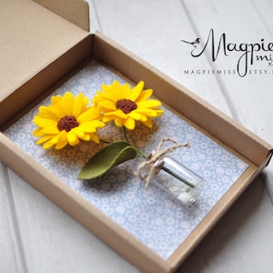 K&ouml;nnte beinhalten: Handgefertigte Geschenkbox mit zwei gelben Filz-Sonnenblumen mit braunen Zentren und einem gr&uuml;nen Filzblatt. Ein kleines Glasfl&auml;schchen mit wei&szlig;en Steinen ist mit Bindfaden gebunden. Die Box ist mit blauem Blumenpapier ausgekleidet.