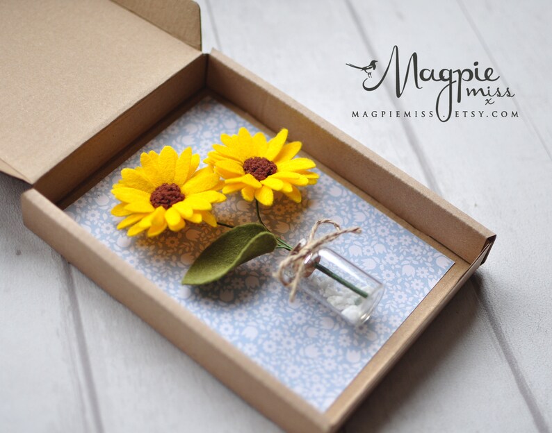 K&ouml;nnte beinhalten: Handgefertigte Geschenkbox mit zwei gelben Filz-Sonnenblumen mit braunen Zentren und einem gr&uuml;nen Filzblatt. Ein kleines Glasfl&auml;schchen mit wei&szlig;en Steinen ist mit Bindfaden gebunden. Die Box ist mit blauem Blumenpapier ausgekleidet.