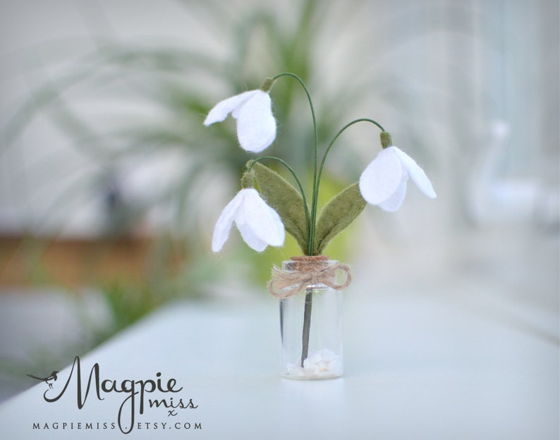 K&ouml;nnte beinhalten: Eine kleine Glasvase mit drei wei&szlig;en Schneegl&ouml;ckchen aus Filz mit gr&uuml;nen Bl&auml;ttern und Stielen. Die Vase ist mit Bindfaden gebunden und steht auf einer wei&szlig;en Oberfl&auml;che. Der Etsy-Shop-Name "Magpie Miss" befindet sich unten links.