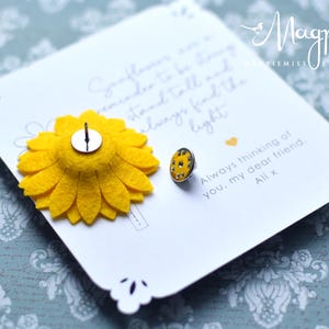 May include: A white card with a yellow felt sunflower pin and a small, round pin with yellow and silver details. The card has a scalloped edge and handwritten text that reads "Always thinking of you, my dear friend, Ali x."