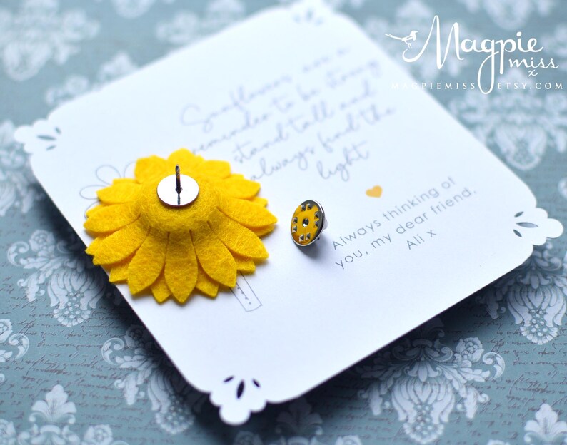 May include: A white card with a yellow felt sunflower pin and a small, round pin with yellow and silver details. The card has a scalloped edge and handwritten text that reads "Always thinking of you, my dear friend, Ali x."