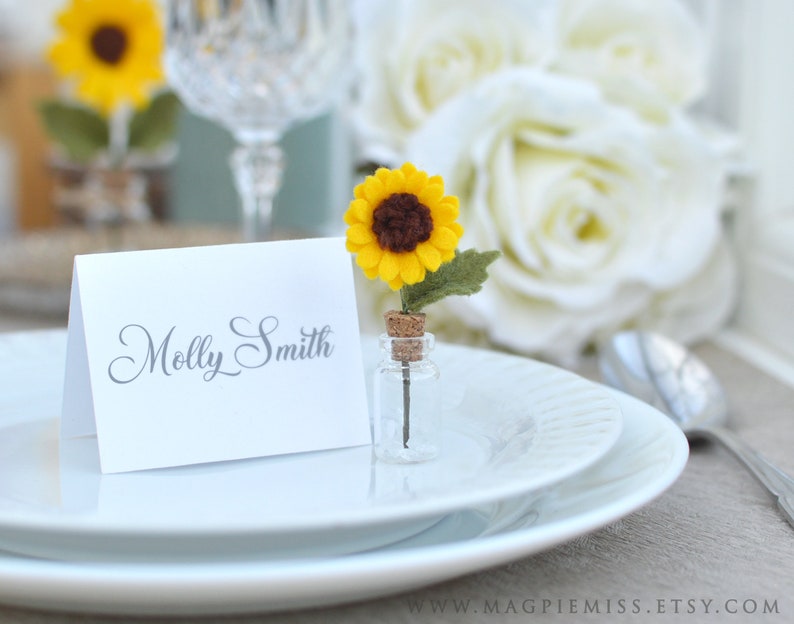 Pu&ograve; includere: Un'impostazione del tavolo con un piatto bianco, un biglietto bianco piegato con il nome "Molly Smith" in corsivo grigio e un girasole in miniatura in una bottiglia di vetro. Il girasole ha petali gialli e un centro marrone.