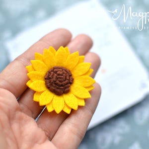 May include: A handmade felt sunflower with bright yellow petals and a brown center. The flower is held in a hand, with a blurred background. The Etsy shop name "MagpieMiss" is visible in the background.
