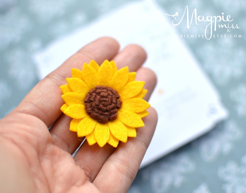May include: A handmade felt sunflower with bright yellow petals and a brown center. The flower is held in a hand, with a blurred background. The Etsy shop name "MagpieMiss" is visible in the background.