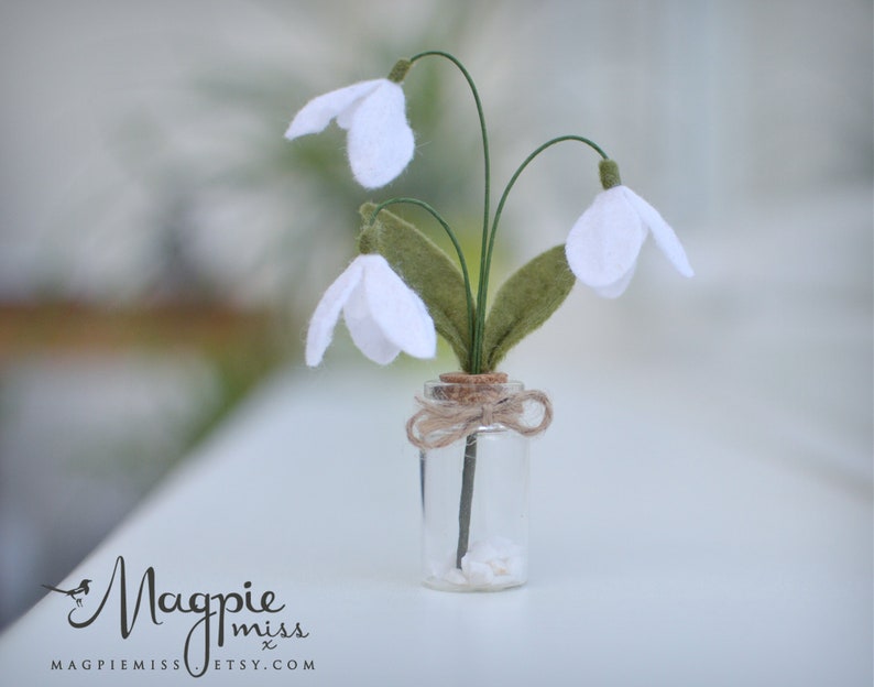 K&ouml;nnte beinhalten: Eine kleine Glasflasche mit einem Korken, gebunden mit Bindfaden, die drei k&uuml;nstliche Schneegl&ouml;ckchenbl&uuml;ten enth&auml;lt. Die Bl&uuml;ten haben wei&szlig;e Bl&uuml;tenbl&auml;tter, gr&uuml;ne Stiele und gr&uuml;ne Bl&auml;tter. Die Flasche enth&auml;lt kleine wei&szlig;e Steine.