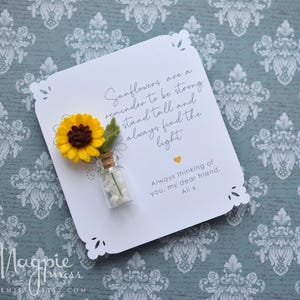 May include: A white card with a decorative edge features a felt sunflower and a small glass bottle. The card includes the text "Sunflowers are a reminder to be strong, stand tall and always find the light."