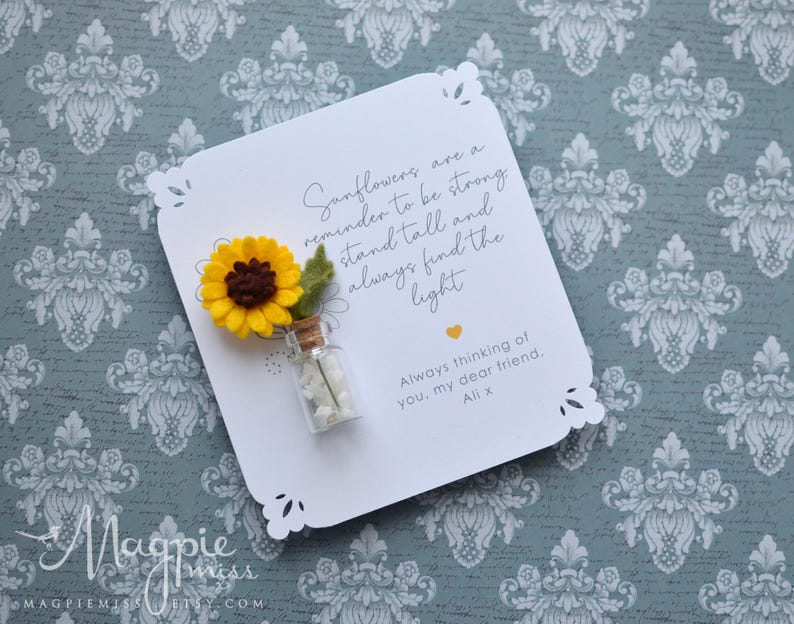 May include: A white card with a decorative edge features a felt sunflower and a small glass bottle. The card includes the text "Sunflowers are a reminder to be strong, stand tall and always find the light."
