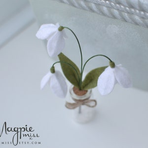 K&ouml;nnte beinhalten: Ein Miniaturarrangement aus Filz-Schneegl&ouml;ckchen in einer kleinen Glasflasche. Die wei&szlig;en Bl&uuml;ten haben gr&uuml;ne Stiele und Bl&auml;tter, und die Flasche ist mit einem Korken verschlossen und mit Bindfaden gebunden. Das Magpie Miss Etsy-Logo ist sichtbar.