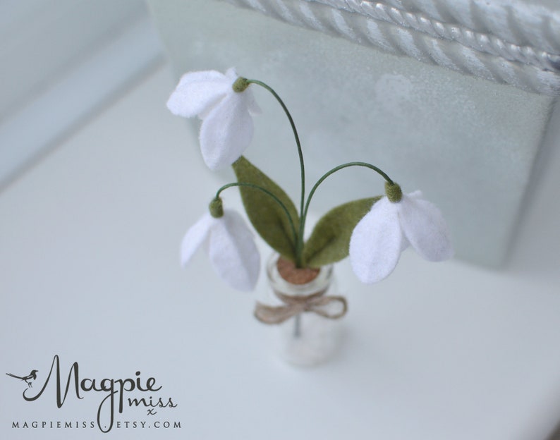 K&ouml;nnte beinhalten: Ein Miniaturarrangement aus Filz-Schneegl&ouml;ckchen in einer kleinen Glasflasche. Die wei&szlig;en Bl&uuml;ten haben gr&uuml;ne Stiele und Bl&auml;tter, und die Flasche ist mit einem Korken verschlossen und mit Bindfaden gebunden. Das Magpie Miss Etsy-Logo ist sichtbar.