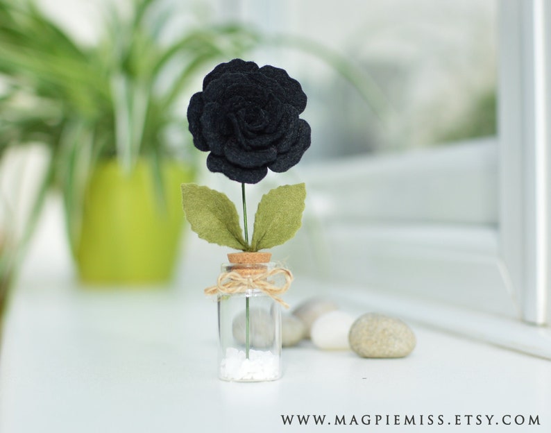 May include: A single black felt rose with green felt leaves in a small glass jar with a cork top and white sand at the bottom.