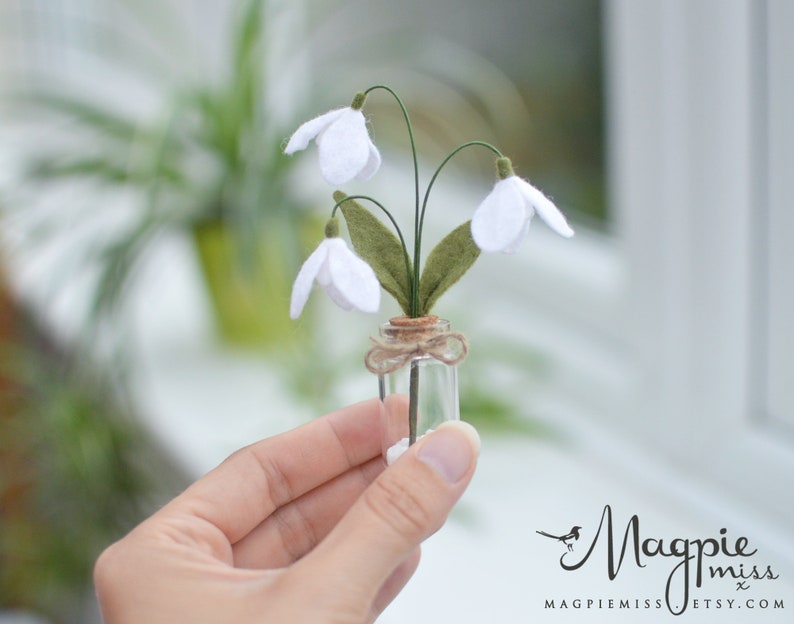 K&ouml;nnte beinhalten: Eine kleine Glasflasche enth&auml;lt ein handgefertigtes Schneegl&ouml;ckchen-Arrangement aus Filz. Die wei&szlig;en Schneegl&ouml;ckchenbl&uuml;ten haben gr&uuml;ne Bl&auml;tter und sind an d&uuml;nnen Drahtstielen befestigt. Die Flasche ist mit Bindfaden gebunden. Das Magpie Miss Etsy-Logo befindet sich unten rechts.
