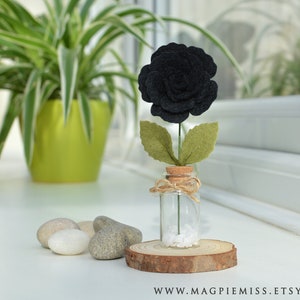 May include: A single black felt rose in a small glass jar with a cork top. The jar is sitting on a wooden slice and there are white stones in the bottom of the jar.