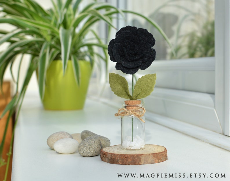 May include: A single black felt rose in a small glass jar with a cork top. The jar is sitting on a wooden slice and there are white stones in the bottom of the jar.