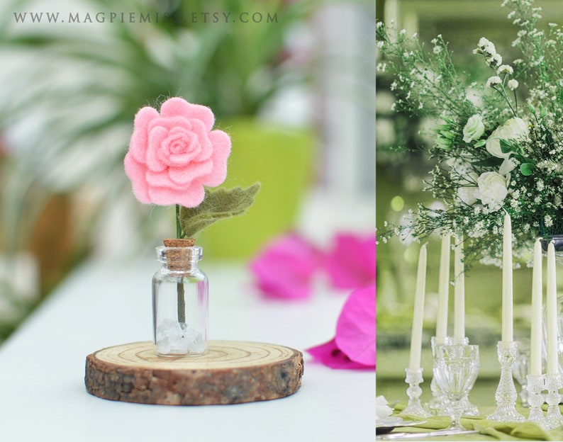 Pu&ograve; includere: Una rosa rosa di feltro con una foglia verde in una piccola bottiglia di vetro con un tappo di sughero, appoggiata su una fetta di legno. Lo sfondo include una composizione floreale sfocata e l'URL del negozio Etsy.