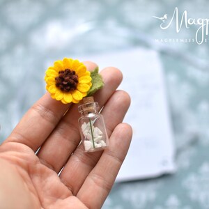 May include: A miniature glass bottle with a cork stopper, filled with white stones, and topped with a felt sunflower. The sunflower has yellow petals, a brown center, and a green leaf. The bottle is held in a person's hand.