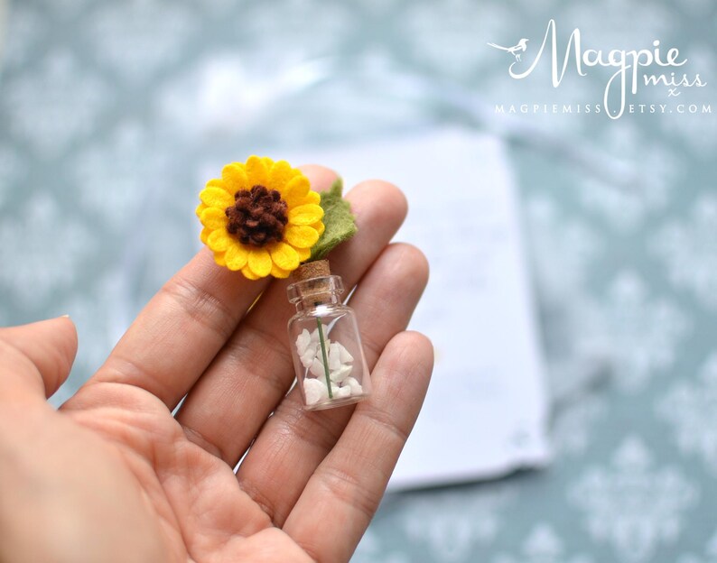 May include: A miniature glass bottle with a cork stopper, filled with white stones, and topped with a felt sunflower. The sunflower has yellow petals, a brown center, and a green leaf. The bottle is held in a person's hand.