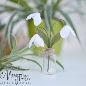 K&ouml;nnte beinhalten: Ein kleines Glasfl&auml;schchen mit Korken enth&auml;lt ein Miniaturarrangement aus wei&szlig;en Schneegl&ouml;ckchenbl&uuml;ten aus Filz. Die Blumen haben gr&uuml;ne Stiele und Bl&auml;tter. Die Flasche ist mit Bindfaden gebunden und steht auf einer wei&szlig;en Oberfl&auml;che.