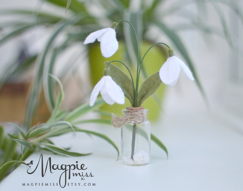 K&ouml;nnte beinhalten: Ein kleines Glasfl&auml;schchen mit Korken enth&auml;lt ein Miniaturarrangement aus wei&szlig;en Schneegl&ouml;ckchenbl&uuml;ten aus Filz. Die Blumen haben gr&uuml;ne Stiele und Bl&auml;tter. Die Flasche ist mit Bindfaden gebunden und steht auf einer wei&szlig;en Oberfl&auml;che.