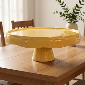 May include: A glossy yellow ceramic cake stand featuring a wide, flat circular top with a slightly raised rim and a sturdy, tapered pedestal base. The smooth, reflective surface of the stand provides a vibrant pop of color for serving.