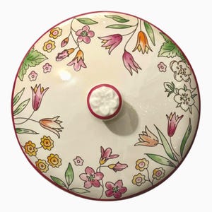 Puede incluir: Una tapa redonda de cerámica con base color crema que presenta un patrón floral pintado a mano con flores rosas, amarillas y naranjas con hojas verdes. La tapa tiene un borde rosa oscuro y un pomo central con forma de pequeña flor blanca.